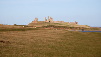 Towards Dunstanburgh This landscape photograph captures the view towards Dunstanburgh Castle in Northumberland, England, showing the historic castle ruins dominating the horizon. Taken late in the morning during the winter season, the grassy foreground reflects the typical colors of winter, while a person can be seen walking towards the castle, adding scale and interest to the scene. Dunstanburgh Castle, managed by English Heritage, stands as a recognizable landmark on the Northumberland coastline in the United Kingdom. The image highlights the contrast between the expansive open land and the impressive medieval fortress, and the presence of people emphasizes its role as a popular destination for visitors.